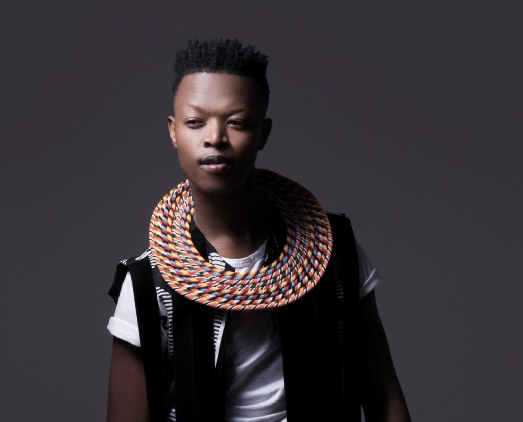 A man looking in to the camera wearing a white tshirt, black waistcoat ND AN AFRICAN TRADITIONAL NECK PIECE