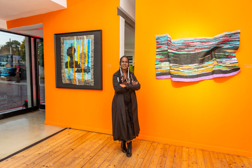A woman stands in front of her artwork and an orange wall