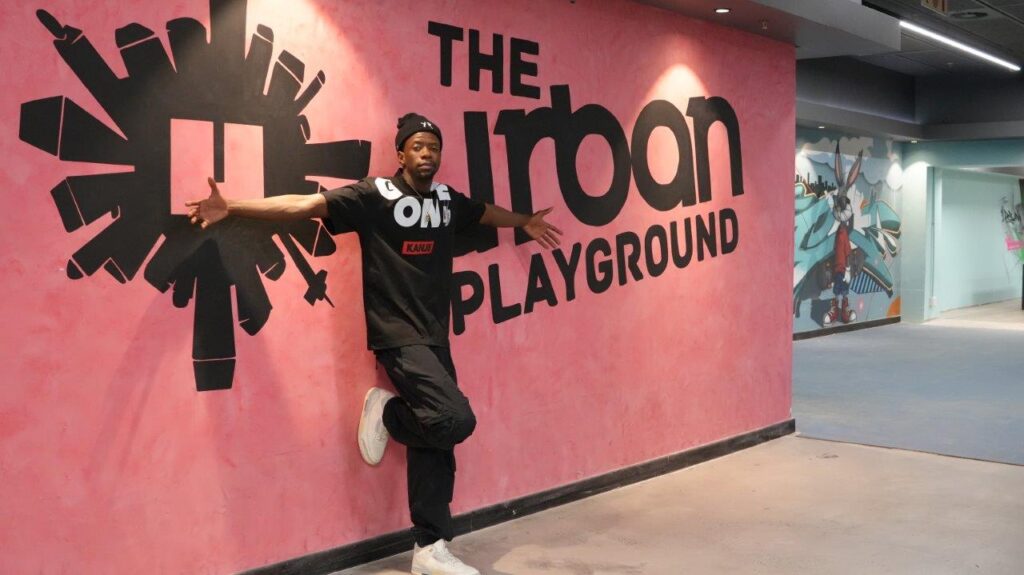 A man poses in front of a pink wall wearing black clothes