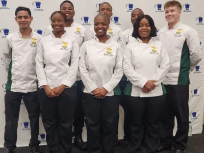 Young chefs dressed in white chef coats stand and pose for the camera in front of the SA Chefs banner