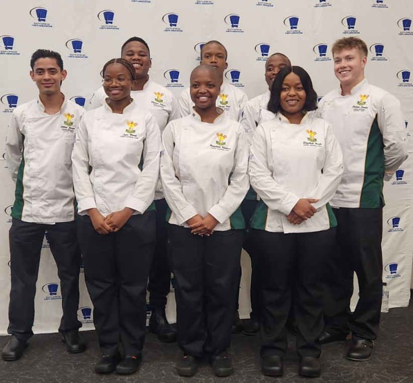 Young chefs dressed in white chef coats stand and pose for the camera in front of the SA Chefs banner