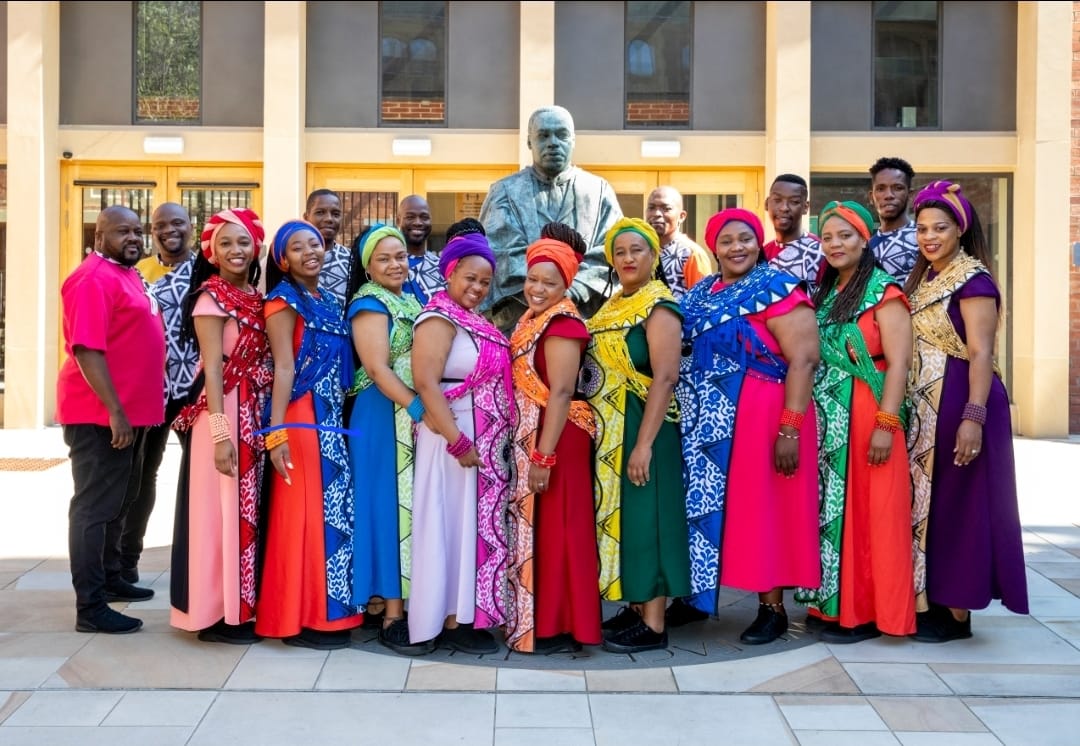 A group of people in a choir pose together wearing colourful clothing in front of a building