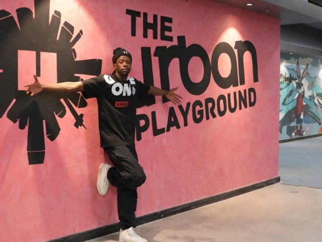 A man poses in front of a pink wall wearing black clothes