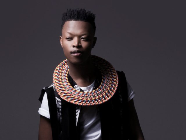 A man looking in to the camera wearing a white tshirt, black waistcoat ND AN AFRICAN TRADITIONAL NECK PIECE