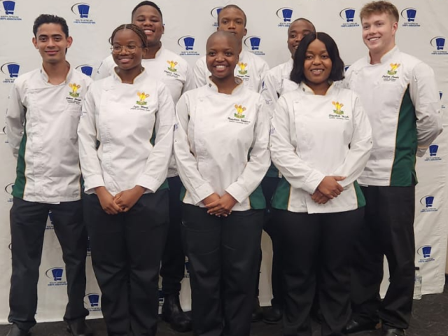 Young chefs dressed in white chef coats stand and pose for the camera in front of the SA Chefs banner