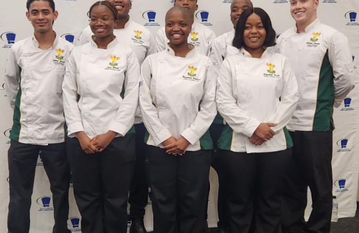Young chefs dressed in white chef coats stand and pose for the camera in front of the SA Chefs banner