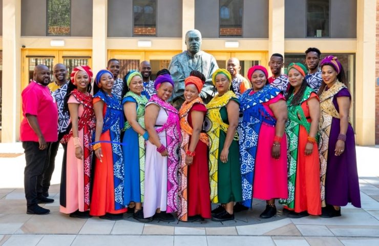 A group of people in a choir pose together wearing colourful clothing in front of a building